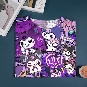 May include: A purple t-shirt featuring cartoon characters with a skull and bow design. The shirt has a graffiti-style background with the word "Gidle" and other text. The shirt is folded on a dark blue surface.