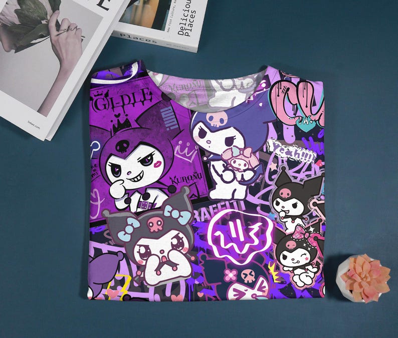 May include: A purple t-shirt featuring cartoon characters with a skull and bow design. The shirt has a graffiti-style background with the word "Gidle" and other text. The shirt is folded on a dark blue surface.