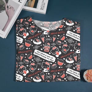 May include: Black t-shirt featuring a repeating pattern of Hello Kitty characters, cupcakes, ice cream cones, and bows in white and red. The shirt includes the text "Hello Kitty" and "You have a way of brightening my day!"