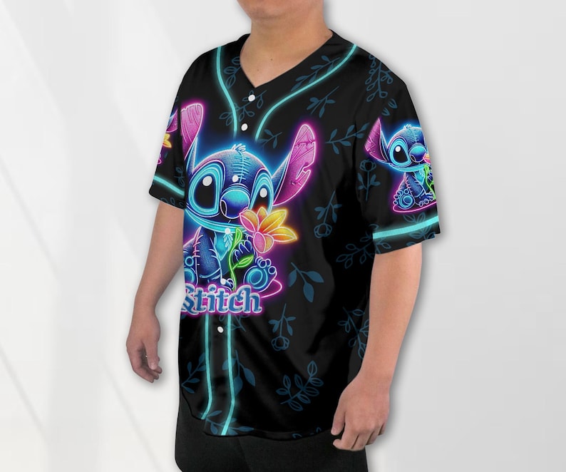 Puede incluir: Una camiseta de b&eacute;isbol negra con un personaje Stitch azul ne&oacute;n sosteniendo una flor. La camiseta tiene un patr&oacute;n floral y la palabra "Stitch" impresa en la parte delantera.