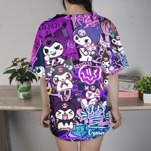 May include: A purple and black graphic t-shirt with cartoon characters and graffiti-style text. The design includes various characters, crowns, and the word "Crown". The shirt has short sleeves and a loose fit.