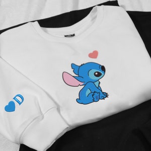May include: White sweatshirt with a blue Stitch character from Lilo & Stitch. Stitch is sitting down and looking up at a pink heart. The sweatshirt has a blue heart with the letter "D" on the sleeve.