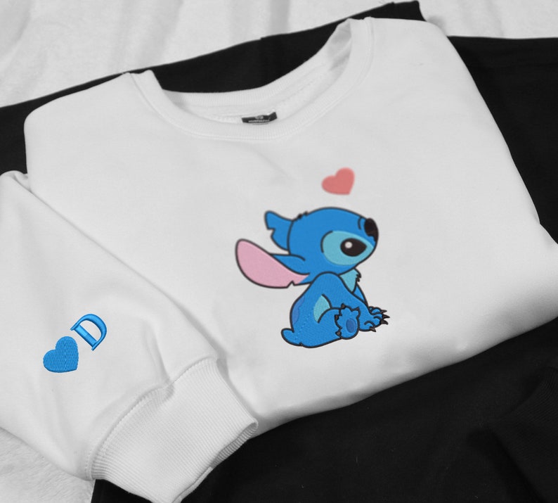 May include: White sweatshirt with a blue Stitch character from Lilo & Stitch. Stitch is sitting down and looking up at a pink heart. The sweatshirt has a blue heart with the letter "D" on the sleeve.