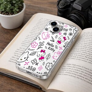 May include: Clear phone case with a Hello Kitty design. The case features a repeating pattern of Hello Kitty characters, flowers, and leaves in black and pink. The words "Hello Kitty" are also printed on the case.