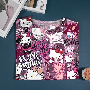 May include: A pink and purple t-shirt featuring multiple illustrations of Hello Kitty with bows and various expressions. The shirt has graffiti-style text including "I Love You" and "ACAB".
