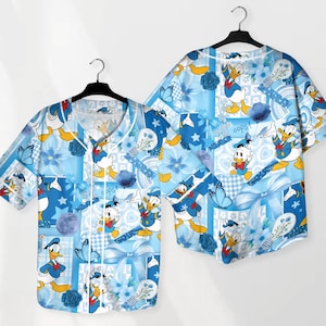 May include: Two short-sleeved button-down shirts with a light blue patchwork design featuring Donald Duck cartoon characters, butterflies, and floral accents. The shirts are hanging on black hangers.