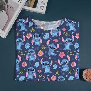 Pu&ograve; includere: Una maglietta blu navy con un motivo ripetuto del personaggio Stitch di Lilo & Stitch. Il design include Stitch, fiori rosa e foglie verdi.