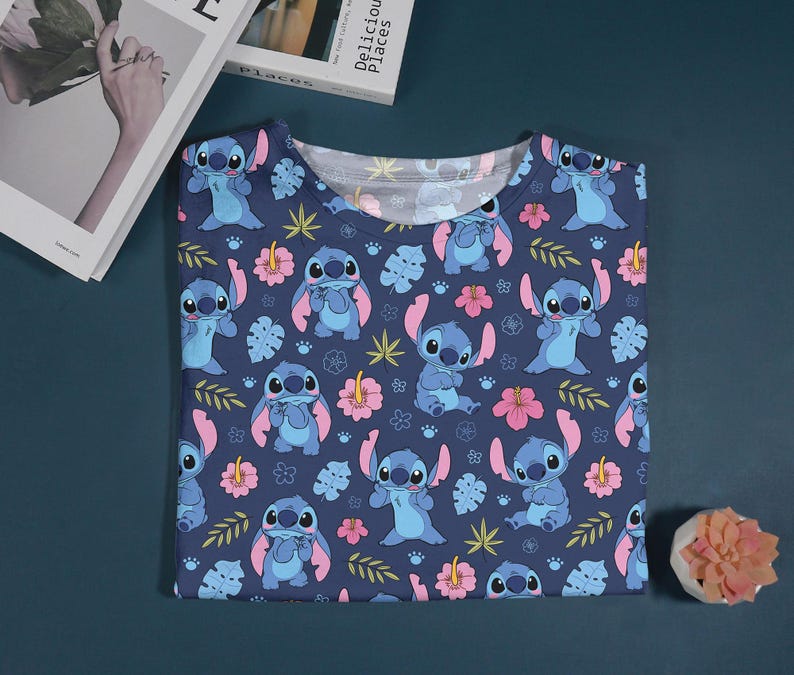 Pu&ograve; includere: Una maglietta blu navy con un motivo ripetuto del personaggio Stitch di Lilo & Stitch. Il design include Stitch, fiori rosa e foglie verdi.