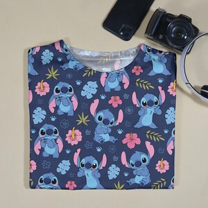 Pu&ograve; includere: Una maglietta blu navy con un motivo ripetuto del personaggio Stitch di Lilo & Stitch, fiori rosa e foglie verdi. La maglietta &egrave; a girocollo.