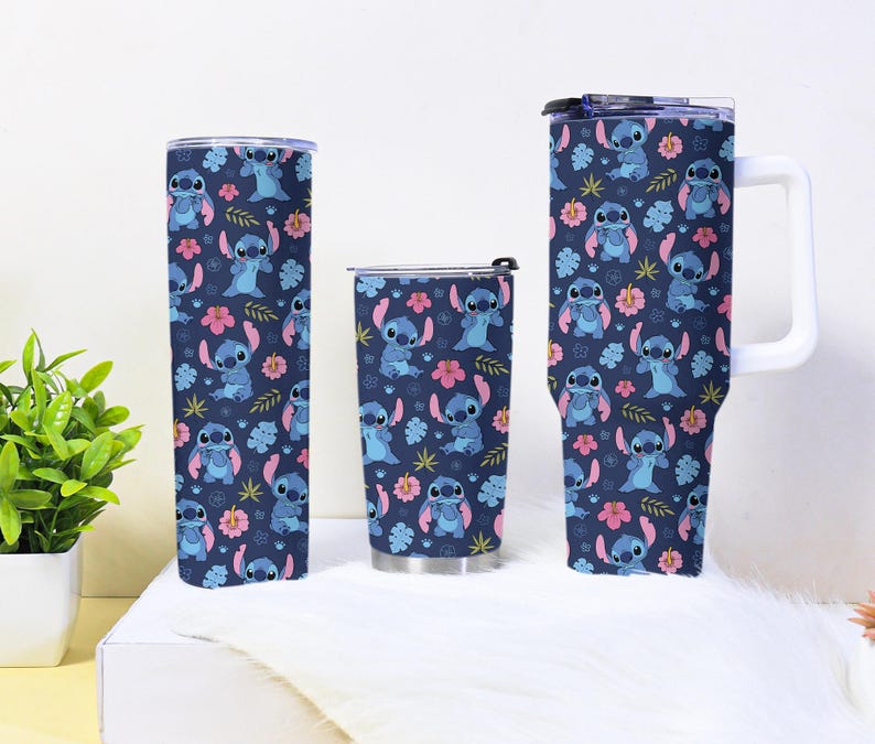 Puede incluir: Tres vasos aislados con un fondo azul marino y un patr&oacute;n repetido de un personaje alien&iacute;gena azul, flores rosas y hojas verdes. Los vasos son de diferentes tama&ntilde;os y formas, incluyendo un vaso alto y delgado, un vaso m&aacute;s corto y una taza grande con un asa blanca.