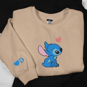 May include: Tan sweatshirt with a blue Stitch character from Lilo & Stitch. Stitch is sitting down and looking up at a pink heart. The sweatshirt has a blue heart with the letter D on the left sleeve.
