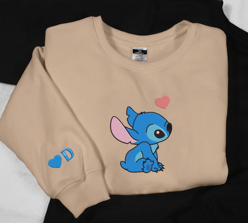 May include: Tan sweatshirt with a blue Stitch character from Lilo & Stitch. Stitch is sitting down and looking up at a pink heart. The sweatshirt has a blue heart with the letter D on the left sleeve.