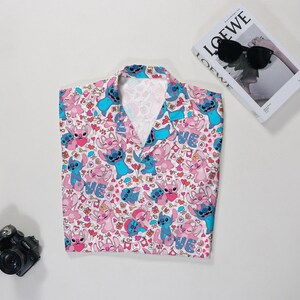 Puede incluir: Una camisa rosa con un estampado de personajes de dibujos animados azules y rosas, corazones y la palabra "LOVE". Tambi&eacute;n se ven una c&aacute;mara negra y un libro con gafas de sol.