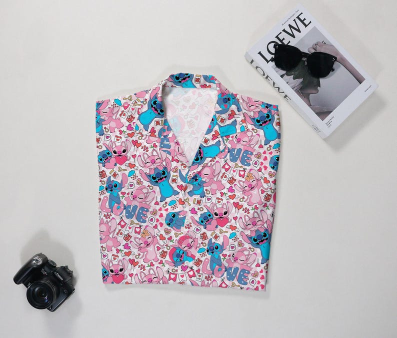 Puede incluir: Una camisa rosa con un estampado de personajes de dibujos animados azules y rosas, corazones y la palabra "LOVE". Tambi&eacute;n se ven una c&aacute;mara negra y un libro con gafas de sol.