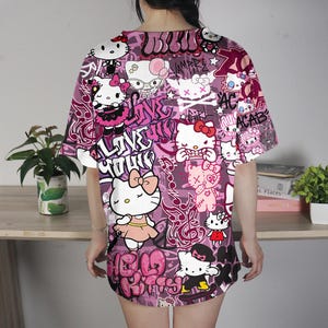 May include: A short-sleeved, oversized t-shirt in shades of pink and purple, featuring a repeating pattern of Hello Kitty characters and graffiti-style text. The design includes various poses of the iconic character, with phrases like "LOVE YOUR" and "ACAB".