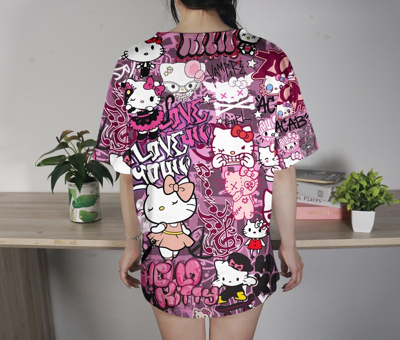 May include: A short-sleeved, oversized t-shirt in shades of pink and purple, featuring a repeating pattern of Hello Kitty characters and graffiti-style text. The design includes various poses of the iconic character, with phrases like "LOVE YOUR" and "ACAB".