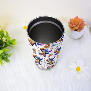 May include: A white tumbler with a colourful pattern of the character Stitch playing basketball. The stainless steel tumbler has a silver rim and interior. The design includes basketball hoops, stars, and Stitch in various poses. The tumbler is on a white surface.