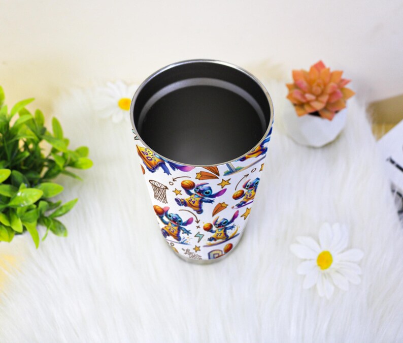 May include: A white tumbler with a colourful pattern of the character Stitch playing basketball. The stainless steel tumbler has a silver rim and interior. The design includes basketball hoops, stars, and Stitch in various poses. The tumbler is on a white surface.