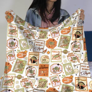 May include: White blanket with a repeating pattern of postage stamp-like designs. The stamps feature cartoon aliens, planets, and phrases like "AMAZE!" and "WHY STUPID?" in orange, green, and brown.