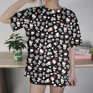 May include: Black t-shirt featuring a repeating pattern of Hello Kitty characters in various Halloween costumes, along with spiderwebs and bats. The design includes the text "Hello Kitty" in white. The shirt has short sleeves.
