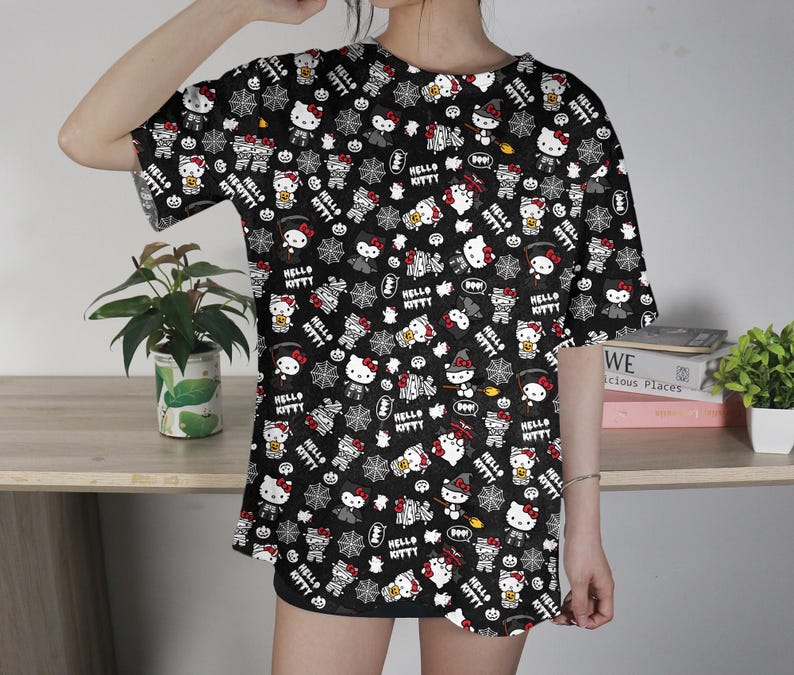 May include: Black t-shirt featuring a repeating pattern of Hello Kitty characters in various Halloween costumes, along with spiderwebs and bats. The design includes the text "Hello Kitty" in white. The shirt has short sleeves.