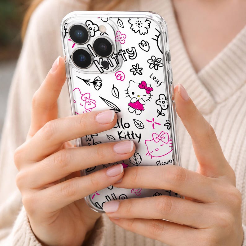 May include: Clear phone case featuring a Hello Kitty design with black and pink illustrations. The case includes drawings of flowers, bows, and the words "Hello Kitty". The case is held in a person's hands.