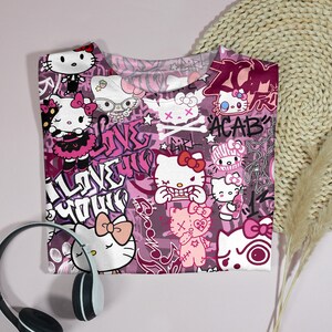 May include: A pink t-shirt featuring a repeating pattern of Hello Kitty characters in various poses, with graffiti-style text and skull and crossbones. The shirt has a crew neck and short sleeves. A pair of headphones is in the foreground.