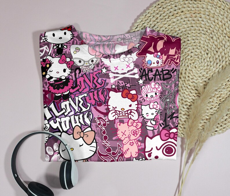 May include: A pink t-shirt featuring a repeating pattern of Hello Kitty characters in various poses, with graffiti-style text and skull and crossbones. The shirt has a crew neck and short sleeves. A pair of headphones is in the foreground.
