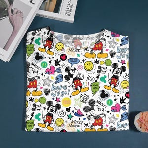 May include: White t-shirt featuring a colorful Mickey Mouse pattern. The design includes Mickey Mouse illustrations, stars, hearts, speech bubbles, and text like "here we go!" and "Mickey Mouse". The shirt is folded on a blue surface.