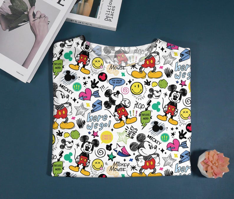 May include: White t-shirt featuring a colorful Mickey Mouse pattern. The design includes Mickey Mouse illustrations, stars, hearts, speech bubbles, and text like "here we go!" and "Mickey Mouse". The shirt is folded on a blue surface.
