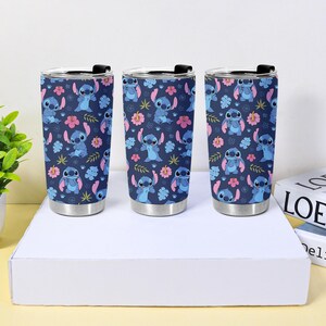 Puede incluir: Tres vasos de acero inoxidable con un patr&oacute;n repetido de Stitch, flores rosas y hojas azules sobre un fondo azul marino. Cada vaso tiene una tapa negra y una base plateada.