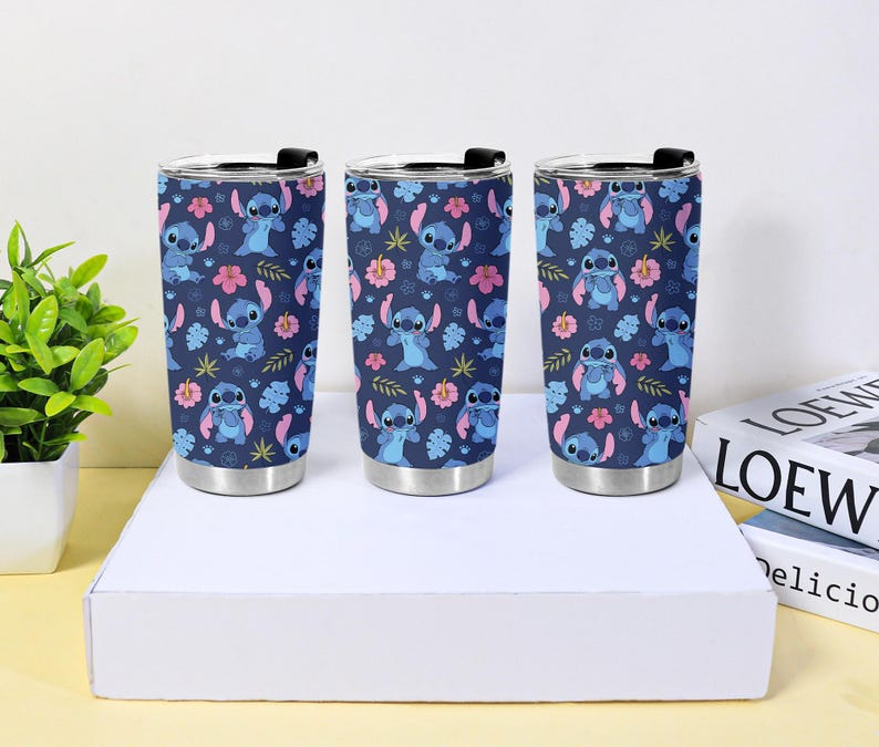 Puede incluir: Tres vasos de acero inoxidable con un patr&oacute;n repetido de Stitch, flores rosas y hojas azules sobre un fondo azul marino. Cada vaso tiene una tapa negra y una base plateada.