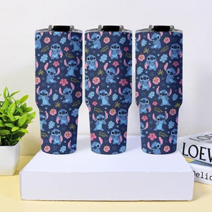 Puede incluir: Tres vasos t&eacute;rmicos azul marino con un patr&oacute;n repetido del personaje Stitch, flores rosas y hojas verdes. Los vasos tienen tapas negras y se exhiben sobre una superficie blanca.