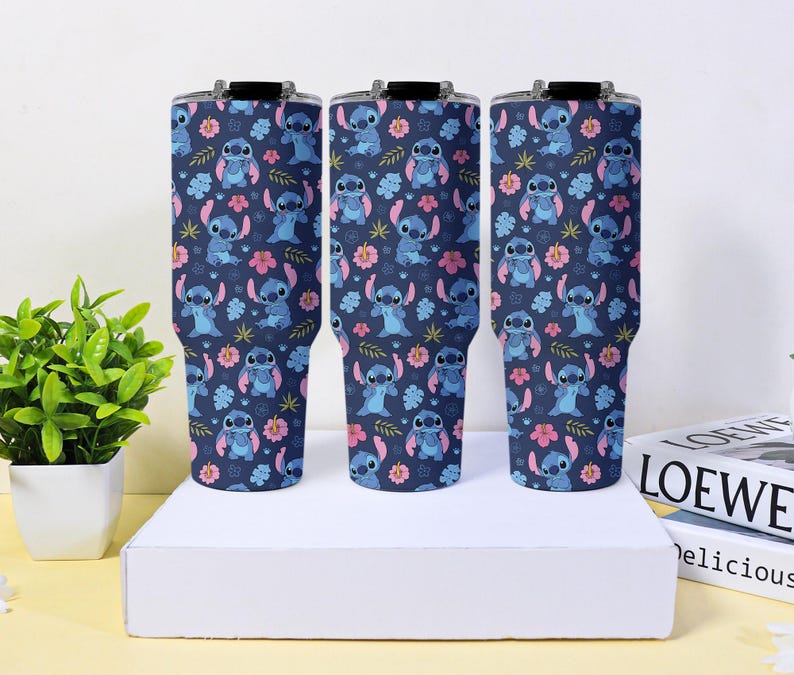 Puede incluir: Tres vasos t&eacute;rmicos azul marino con un patr&oacute;n repetido del personaje Stitch, flores rosas y hojas verdes. Los vasos tienen tapas negras y se exhiben sobre una superficie blanca.