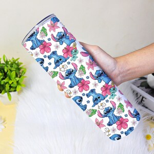 Puede incluir: Un vaso blanco con un patr&oacute;n repetido del alien&iacute;gena azul Stitch, flores rosas y hojas verdes. El vaso est&aacute; sostenido por la mano de una persona.
