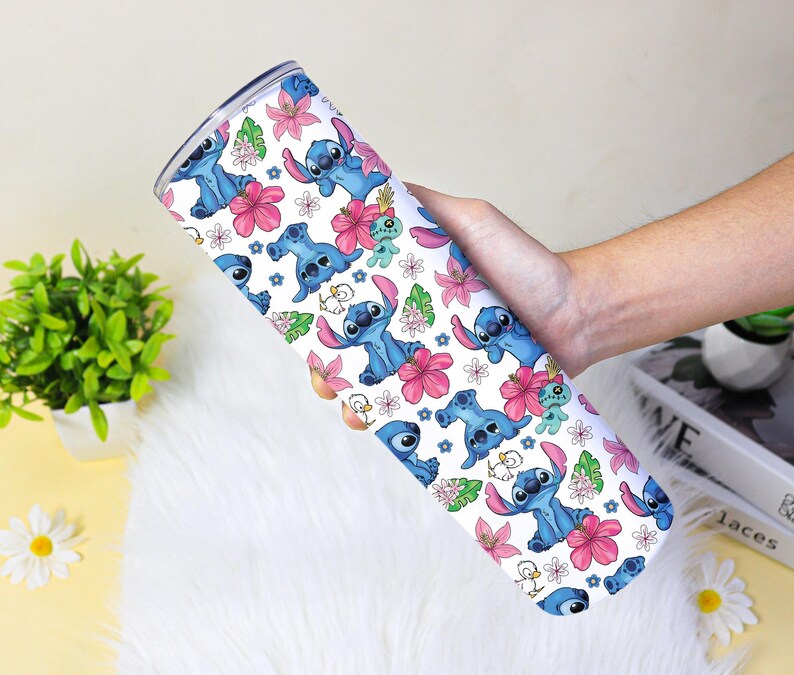 Puede incluir: Un vaso blanco con un patr&oacute;n repetido del alien&iacute;gena azul Stitch, flores rosas y hojas verdes. El vaso est&aacute; sostenido por la mano de una persona.