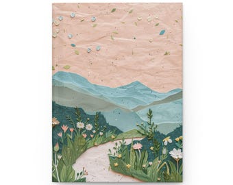 May include: A hardcover journal featuring a landscape illustration. The cover art depicts a path winding through a field of wildflowers, leading towards layered blue mountains under a pink sky. Decorative paper elements add texture and depth to the design.