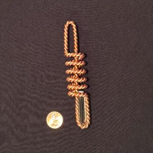 以下が含まれることがあります： 手作りの銅製ペンダント。巻き付けられたデザインで、長方形の濃い緑色のクリスタルが特徴です。 サイズの目安として、ペンダントの隣にペニーが写っています。 銅は暖かく赤みがかった茶色です。