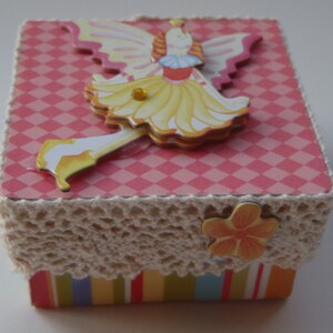 Puede incluir: Una pequeña caja rosa y blanca con un borde de encaje decorativo. La caja presenta un patrón de rayas coloridas en la parte inferior y una imagen 3D de un hada con alas y un vestido amarillo.