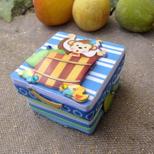 May include: A small blue and white striped box with a monkey holding a bucket and a yellow duck on the lid. The box has a gold handle and a green button.