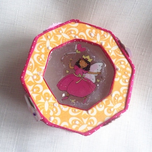 Puede incluir: Una caja octogonal amarilla y rosa con una ilustración de hada blanca y rosa en su interior. La caja tiene un diseño estampado blanco y amarillo con acentos de perlas blancas.