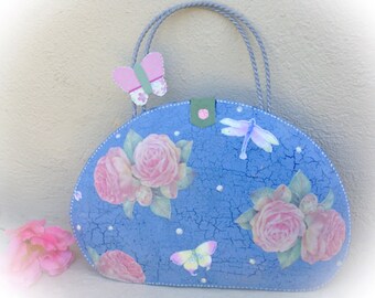 Può includere: Una borsa blu con un design floreale con rose rosa e farfalle. La borsa ha una texture screpolata, un manico grigio e una farfalla decorativa sul lato. Un fiore rosa è accanto alla borsa.