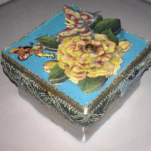 Puede incluir: Caja decorativa cuadrada con una tapa azul claro brillante, adornada con una gran flor amarilla y mariposas. La caja tiene un borde plateado y dorado, y una base de cinta plateada estampada. El diseño es ornamentado y detallado.