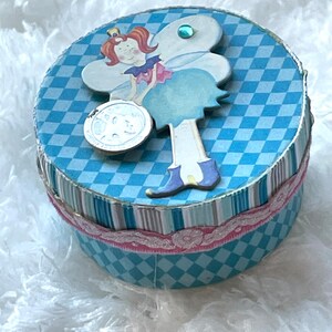 Puede incluir: Una caja redonda azul y blanca a cuadros con una cinta rosa y una figura de hada en la tapa. El hada lleva un vestido azul, tiene el pelo rojo y una corona.