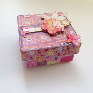 Puede incluir: Una pequeña caja cuadrada con un patrón floral rosa, morado y amarillo. La caja tiene un recorte de papel 3D de una bailarina con alas rosas y blancas y un vestido rosa. La caja está decorada con una pequeña mariposa rosa.