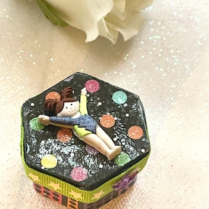 Puede incluir: Una pequeña caja hexagonal con fondo negro y lunares de colores. Una figura de una gimnasta está unida a la parte superior de la caja. La caja está decorada con una cinta verde, negra y blanca.