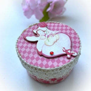 Puede incluir: Una pequeña caja de cuadros rosa y blancos con un borde de encaje blanco. La caja tiene un recorte de madera decorativo de un hada con una flor sobre un fondo rosa.
