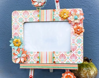 May include: A white picture frame with an easel stand, decorated with orange, yellow, and teal flowers. The frame has a patterned background with a damask design.
