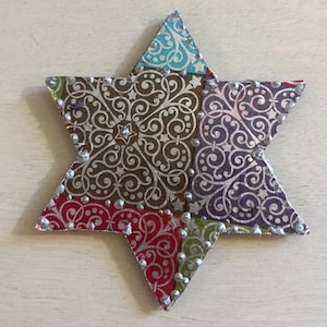 Peut inclure: Une étoile à six branches ornementale composée de sections en patchwork aux couleurs variées, notamment marron, violet, rouge, vert et bleu. Chaque section présente un motif filigrane argenté et est bordée de points argentés en relief.