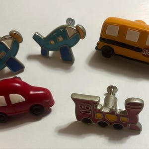 May include: A set of five decorative cabinet knobs shaped like vehicles, including two blue airplanes, a yellow school bus with a red stop sign, a red car with white windows, and a red and silver train engine with a metal mounting screw.